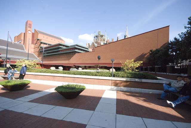 The red brick bulk of the British Library can be seen across a paved patterned countyard planted with shrubs and tubs. The spires of the St Pancras hotel can be seen in the background.