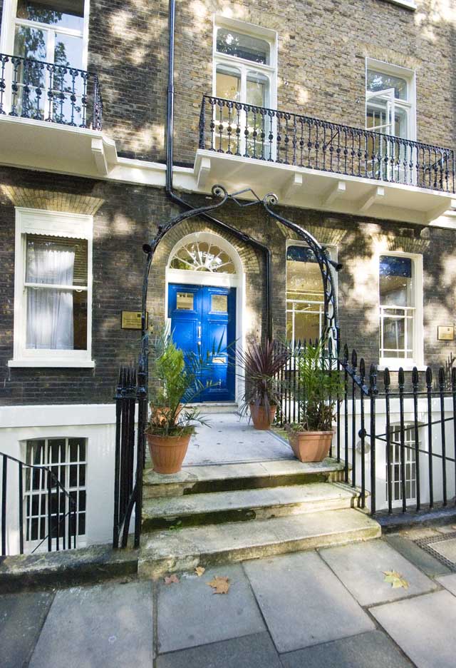 The Georgian house is up steps which are framed with a wrought iron lamp stand. The steps lead up to a blue double door with an elegant fan light above it and sash windows to either side.